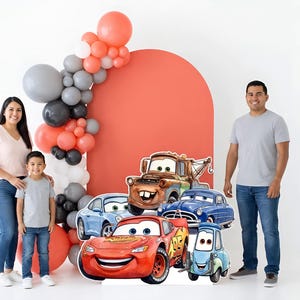 May include: A family poses with a Cars-themed backdrop featuring cardboard cutouts of Lightning McQueen, Mater, and other characters. A balloon arch in coral, gray, and black adds to the festive scene.