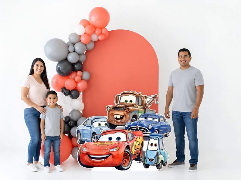 May include: A family poses with a Cars-themed backdrop featuring cardboard cutouts of Lightning McQueen, Mater, and other characters. A balloon arch in coral, gray, and black adds to the festive scene.