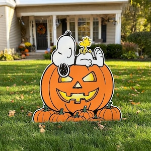 May include: A flat outdoor yard sign featuring a large orange jack-o'-lantern with a glowing face, topped with a white Snoopy dog and a small yellow Woodstock bird. Several smaller orange pumpkins are arranged at the base of the large pumpkin.