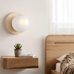 May include: A modern bedroom scene featuring a wall-mounted light fixture with a cream-colored base and a spherical, frosted glass shade. A small wooden nightstand holds a potted plant and a book. A wooden bed frame and a bed with a blanket are also visible.