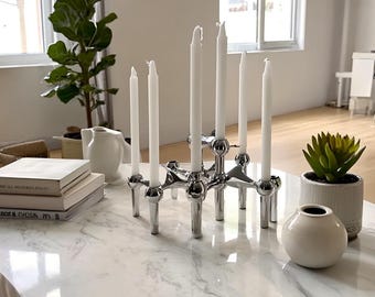 Puede incluir: Escena de salón moderno con un candelabro plateado con velas blancas. Una pila de libros, un pequeño jarrón blanco y una suculenta en maceta están sobre una mesa de centro con tapa de mármol. También se ve una jarra blanca.