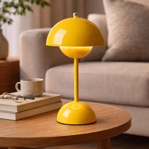 May include: A bright yellow table lamp with a rounded shade and base, and a slender stem. The lamp has a silver-colored top and is placed on a wooden table. A cup, glasses, and books are also on the table.