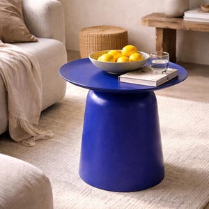 Puede incluir: Una mesa de centro azul vibrante, con una parte superior redonda y una base cilíndrica. La mesa sostiene un cuenco de limones, un libro y un vaso de agua. La mesa está sobre una alfombra de color claro. Mide unos 45 cm de alto.