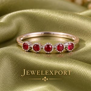 Könnte beinhalten: Ein zarter Ring mit fünf runden, leuchtend roten Rubinen, die in eine polierte Metallschiene gefasst sind. Jeder Edelstein ist mit einer kleinen, perlenartigen Krappenfassung gesichert, wodurch eine durchgehende Reihe auf dem Ring entsteht. Der Text JEWELEXPORT ist sichtbar.