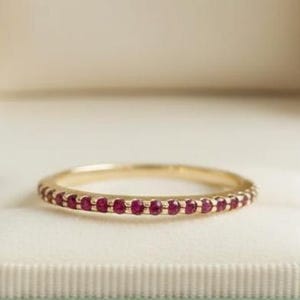 May include: A gold band ring with small, red gemstones set around the entire band. The ring is displayed inside a light green jewelry box with a cream-colored interior.