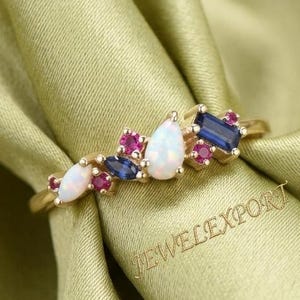 Könnte beinhalten: Ein Goldring mit einer Ansammlung von Edelsteinen, darunter zwei tropfenförmige Opale, zwei marquise-geschliffene blaue Saphire, ein baguette-geschliffener blauer Saphir und mehrere kleine runde rosa Rubine. Der Text JEWELEXPORT ist sichtbar.
