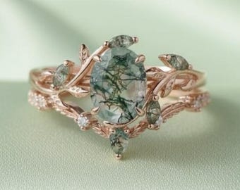May include: A rose gold ring set featuring an oval-cut moss agate center stone. The ring is accented with delicate leaf and vine details, and small clear stones. The set is displayed on a light green, silky fabric.