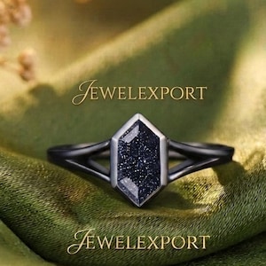 May include: A black metal ring featuring a hexagonal-cut dark blue sandstone with sparkling inclusions. The band has a split-shank design that meets at the central stone. The text JEWELEXPORT is visible above and below the ring in gold lettering.