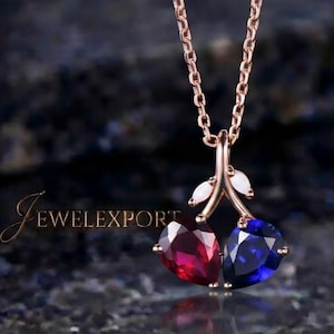 May include: A rose gold necklace featuring a pendant shaped like two cherries, with one heart-shaped red gemstone and one heart-shaped blue gemstone, accented by two small white leaves. The text JEWELEXPORT is visible on the left side.