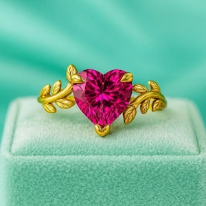 May include: A gold ring with a heart-shaped, magenta gemstone. The ring features a leaf design on the band. The ring is displayed on a green velvet box against a teal background.