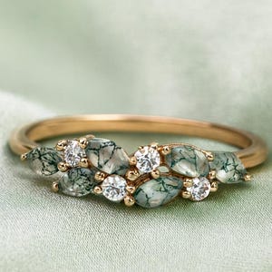 Puede incluir: Un anillo de oro con piedras de ágata musgosa verde y diamantes blancos brillantes. El diseño del anillo se asemeja a un delicado arreglo floral, con las piedras preciosas dispuestas en un patrón de hojas. El anillo se muestra sobre una tela verde suave.