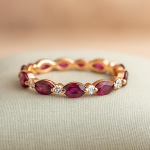 May include: A gold ring featuring alternating marquise-cut rubies and round diamonds. The ring's band is a warm gold color, and the rubies are a deep red. The diamonds add a touch of sparkle to the design. The ring is set on a light green surface.