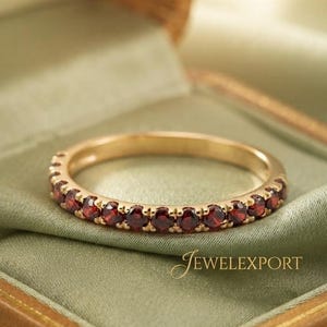 May include: A thin gold band ring featuring a continuous row of small, round, deep red garnet gemstones set in a prong setting. The ring is positioned horizontally, and the text JEWELEXPORT is visible in the lower right corner of the image.