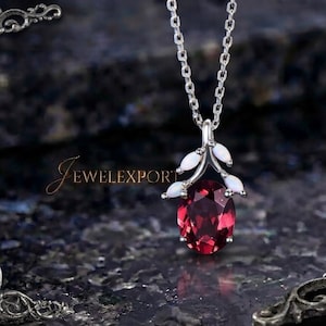 May include: A silver-toned necklace featuring an oval-cut deep red gemstone pendant suspended from a delicate chain. The pendant is accented with four small, white, marquise-shaped stones arranged in a branch-like design above the red gem.