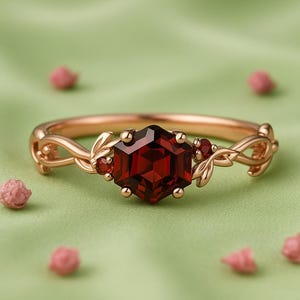 May include: A rose gold ring with a large, hexagonal, deep red gemstone as the focal point. The ring features smaller red gemstones and leaf-like details on the band. The ring is set against a soft green background.