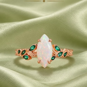 Könnte beinhalten: Ein Ring aus Roségold mit einem zentralen, marquise-geschliffenen Opal und kleinen, marquise-geschliffenen grünen Steinen in einem blattartigen Muster entlang der Ringschiene. Der Text JEWELEXPORT ist oben im Bild zu sehen.