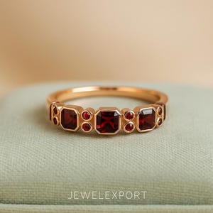 May include: A gold ring featuring a series of square and round red gemstones. The ring has a delicate band and is set against a soft, neutral background. The text "JEWELEXPORT" is visible at the bottom.