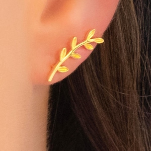 Puede incluir: Un pendiente trepador dorado con forma de delicada rama con hojas. El pendiente se lleva en la oreja, mostrando su intrincado diseño y acabado brillante. El pendiente es un accesorio elegante.