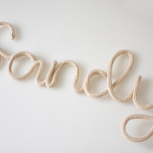 K&ouml;nnte beinhalten: Der Name "Sandy" aus beigefarbenem Strickgarn in Schreibschrift. Die Buchstaben sind verbunden und scheinen aus dickem, weichem Garn zu bestehen. Der Name wird auf wei&szlig;em Hintergrund angezeigt.