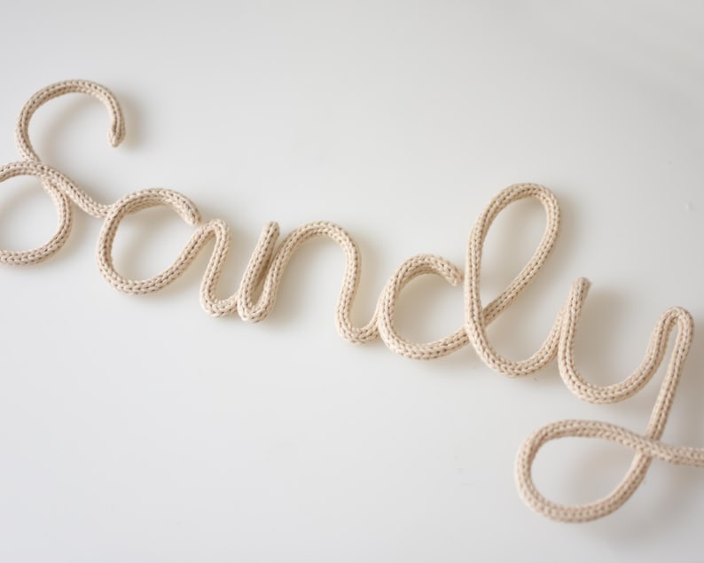 K&ouml;nnte beinhalten: Der Name "Sandy" aus beigefarbenem Strickgarn in Schreibschrift. Die Buchstaben sind verbunden und scheinen aus dickem, weichem Garn zu bestehen. Der Name wird auf wei&szlig;em Hintergrund angezeigt.