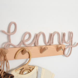 K&ouml;nnte beinhalten: Eine hellbraune Wandgarderobe mit drei Haken. Das Wort "Lenny" ist in einem kursiven, gestrickten, hellrosa Garn geschrieben. Ein Kleiderb&uuml;gel aus Rattan und Babykleidung h&auml;ngen darunter.