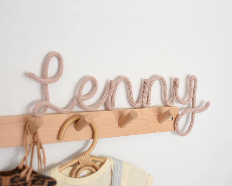 K&ouml;nnte beinhalten: Eine hellbraune Wandgarderobe mit drei Haken. Das Wort "Lenny" ist in einem kursiven, gestrickten, hellrosa Garn geschrieben. Ein Kleiderb&uuml;gel aus Rattan und Babykleidung h&auml;ngen darunter.