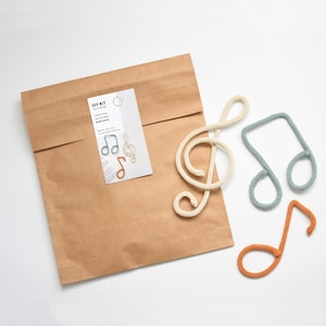 Puede incluir: Un sobre de papel marrón con una etiqueta blanca que dice "DIY KIT - Crea tus propias notas musicales en miniatura". Hay cuatro notas musicales de ganchillo en una superficie blanca: una clave de sol, una negra, una blanca y una semicorchea.