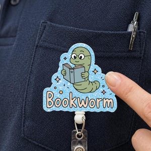 Könnte beinhalten: Ein blau-weißes "Bookworm"-Ausweis-Jojo mit einem Cartoon-Wurm, der eine Brille trägt und ein Buch liest. Das Ausweis-Jojo ist an einem marineblauen Poloshirt mit einer Tasche und dem Logo "UPBUD GROCERY" befestigt.