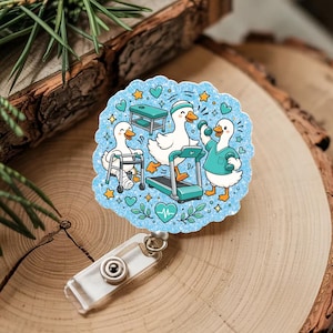 Puede incluir: Un portatarjetas circular con fondo de purpurina azul claro y una ilustración de tres patos blancos. Un pato usa un andador, otro corre en una cinta de correr y el tercero levanta pesas. El diseño incluye pequeños corazones, estrellas y hojas.