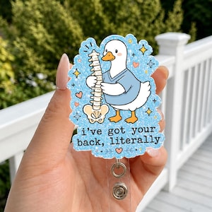 Puede incluir: Un portatarjetas con un pato de dibujos animados sosteniendo una columna vertebral, con el texto "I've got your back, literally". El pato lleva una camisa azul. El portatarjetas tiene una base de plástico transparente y un cordón retráctil.