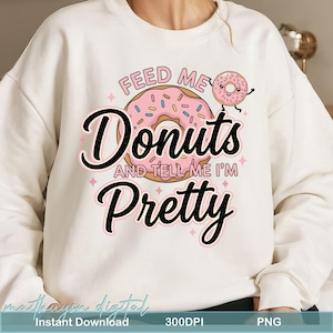 May include: White sweatshirt with a pink donut graphic and the text "Feed Me Donuts And Tell Me I'm Pretty". The donut has pink frosting and colorful sprinkles. The sweatshirt also has a small donut character.