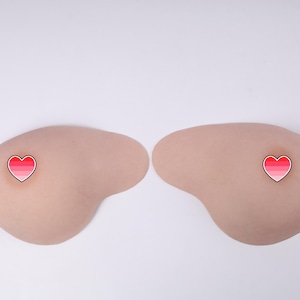 Op de afbeelding: Twee huidkleurige borstvormen met hartvormige tepelstickers. De hartstickers hebben een rode en roze gradiënt met een witte rand. De achtergrond is wit.