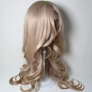 May include: A light blonde wig with long, wavy curls. The wig is styled on a white wig stand against a white background. The hair has a natural-looking color and texture, with soft waves and curls.