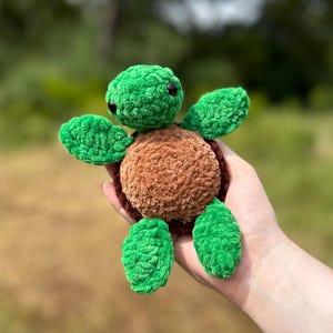 May include: A handmade crochet turtle in shades of green and brown. The turtle has a green head, flippers, and feet, with a brown shell. The turtle is a soft, plush toy.
