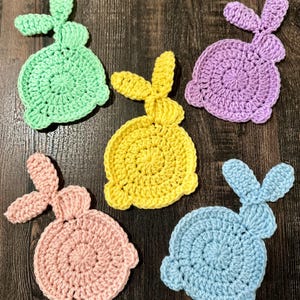May include: Five handmade crochet bunny coasters in pastel colors: green, purple, yellow, pink, and blue. Each coaster features a circular body and two upright ears, perfect for Easter or spring decor.