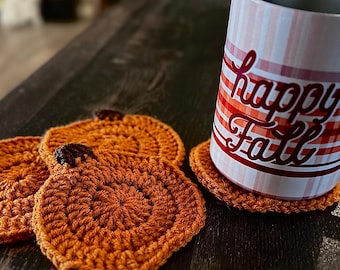 Puede incluir: Posavasos de calabaza naranja tejidos a mano con tallos marrones. Un vaso con un estampado de cuadros rojos y blancos y la inscripción "happy Fall" está colocado sobre uno de ellos. Decoración otoñal.