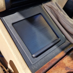Op de afbeelding: Auto-interieurconsole met een zwarte schuifklep, beige leer en houten afwerking. De console heeft een rechthoekig opbergvak en een bekerhouder. Een veiligheidsgordelgesp is zichtbaar.