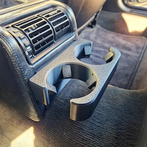 May include: A black, 3D-printed car cup holder with two circular openings. The cup holder is mounted on a black leather car interior, next to an air vent. The car's interior is a dark gray color.