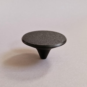 Peut inclure: Bouton de meuble noir en forme de champignon, avec un dessus texturé et moucheté. Le bouton a un dessus plat et arrondi et une base conique. Ce matériel est conçu pour les tiroirs ou les portes d'armoires.