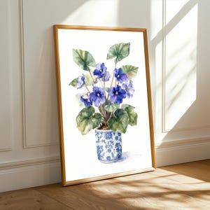 May include: A framed watercolour painting of an African violet plant. The plant has purple flowers and green leaves. The pot is blue and white ceramic with a floral design. The frame is made of wood.