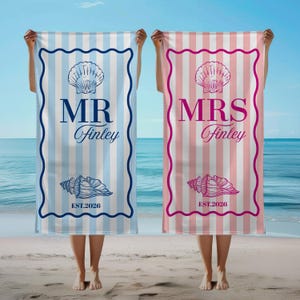 Può includere: Due teli da mare, uno azzurro chiaro e uno rosa, con righe verticali e disegni di conchiglie. Il telo blu dice "MR Finley EST.2026" e quello rosa "MRS Finley EST.2026".