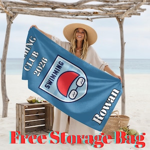 Puede incluir: Una toalla de natación azul con el texto "Swimming Club 2026" y "Rowan". La toalla presenta un logotipo con temática de natación con un gorro de baño rojo y gafas. La toalla se sostiene en una playa.
