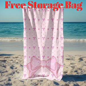 Puede incluir: Una toalla de playa rosa con un gran diseño de lazo y el nombre "Seraphina" impreso. La toalla tiene un patrón de pequeños lazos y flores. Las palabras "Free Storage Bag" están en la parte superior. La toalla está colgada en una playa.