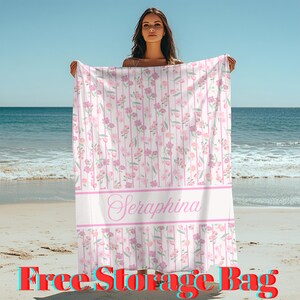 May include: A beach towel with a floral pattern in shades of pink and white, with vertical stripes. The towel has the name "Seraphina" in a cursive font. The words "Free Storage Bag" are at the bottom.
