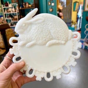 May include: A white, decorative plate featuring a detailed relief of a rabbit. The plate has a scalloped edge with a series of looped and clover-shaped cutouts. The rabbit is positioned above a circular area, and the overall design is vintage-inspired.