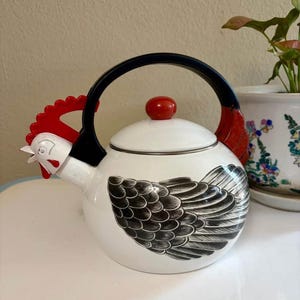 May include: White ceramic teapot with a black and white wing design. The teapot features a red rooster head and tail, a black handle, and a red knob on the lid. The teapot is designed to resemble a chicken.