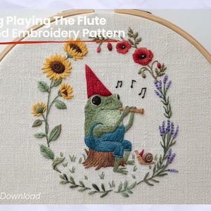 Op de afbeelding: Handgeborduurd patroon met een kikker die fluit speelt, met een rode hoed en blauwe tuinbroek, omringd door zonnebloemen, klaprozen en lavendel. De tekst "Frog Playing The Flute Hand Embroidery Pattern" staat bovenaan.