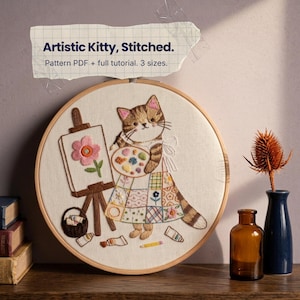 Può includere: Un telaio da ricamo rotondo in legno contenente un pannello in tessuto con l'illustrazione ricamata di un gatto soriano marrone con grembiule patchwork, che tiene una tavolozza, accanto a un cavalletto con un fiore e il testo Artistic Kitty, Stitched.