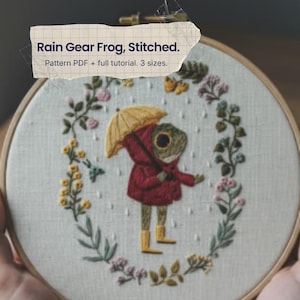 May include: A circular embroidery hoop containing a fabric piece with a stitched frog wearing a red raincoat and yellow boots, holding a yellow umbrella. The frog is surrounded by a floral wreath and rain drops. Text reads: Rain Gear Frog, Stitched. Pattern PDF + full tutorial. 3 sizes.