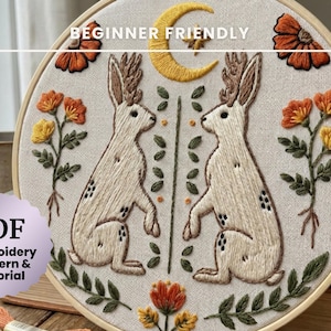 Könnte beinhalten: Gesticktes Wandbild in einem Holzrahmen mit zwei Hasen, einem Mond und Sternen. Das Design beinhaltet orangefarbene und gelbe Blumen. Der Text lautet "BEGINNER FRIENDLY" und "PDF Embroidery Pattern & Tutorial".